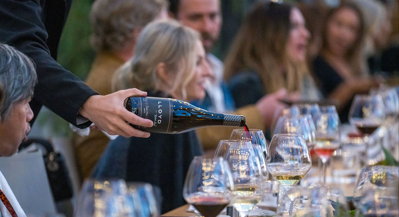 A photo of a glass of Pinot Noir being poured to guests with host Bonnie Lloyd seated in the background.