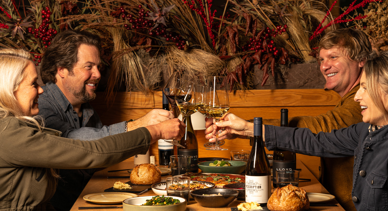 Friends raising glasses of Prescription Chardonnay around a festive holiday table set with shared dishes, bread, and wine bottles.