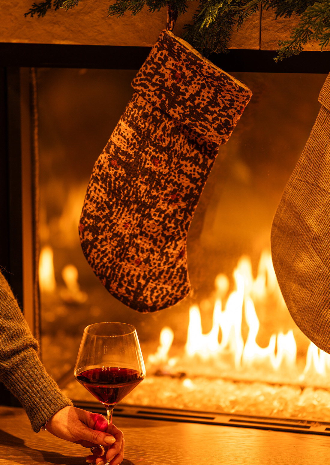 Hand holding a glass of red wine in front of a glowing fireplace with hanging holiday stockings and greenery.”