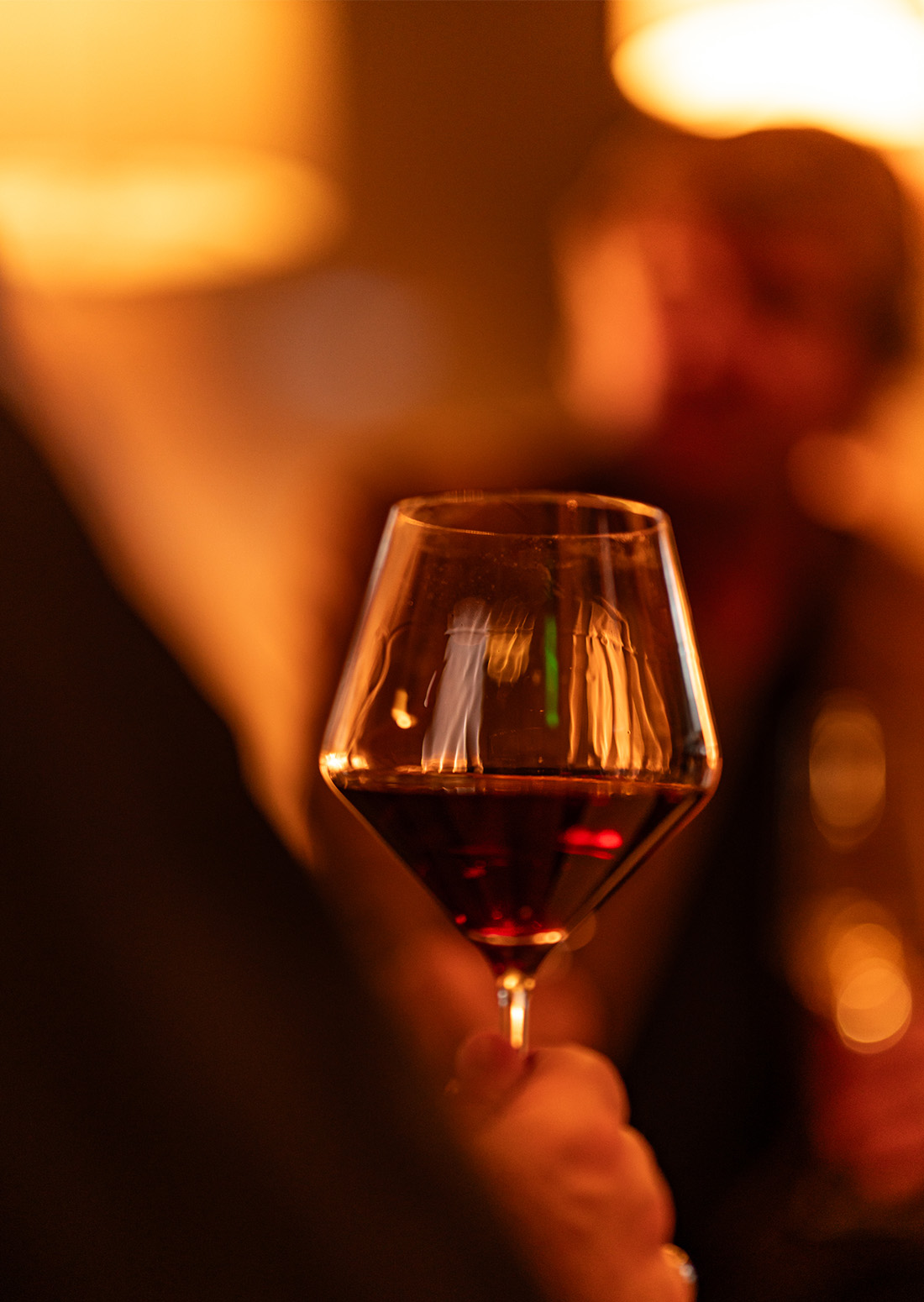Close-up of a hand holding a glass of red wine in warm, ambient light during an intimate gathering.