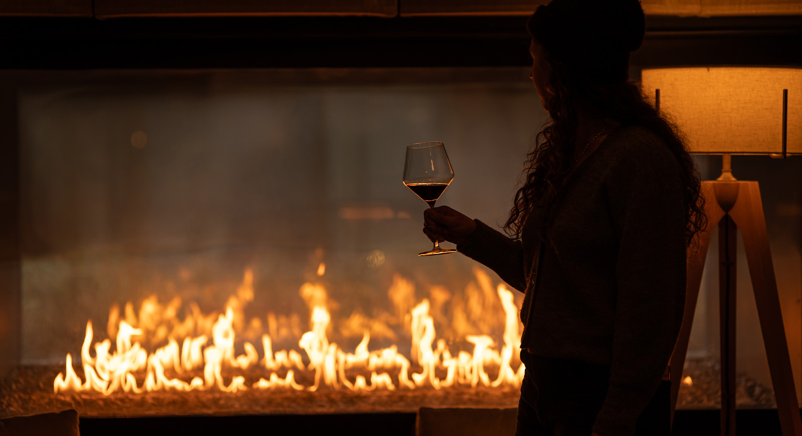Silhouette of a woman holding a glass of red wine while standing by a glowing fireplace in warm, ambient light.