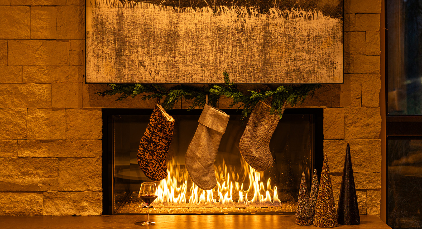A warm holiday fireplace scene with three neutral-toned stockings hanging above the fire, evergreen garland, and a glass of red wine on the hearth at Lloyd Cellars.