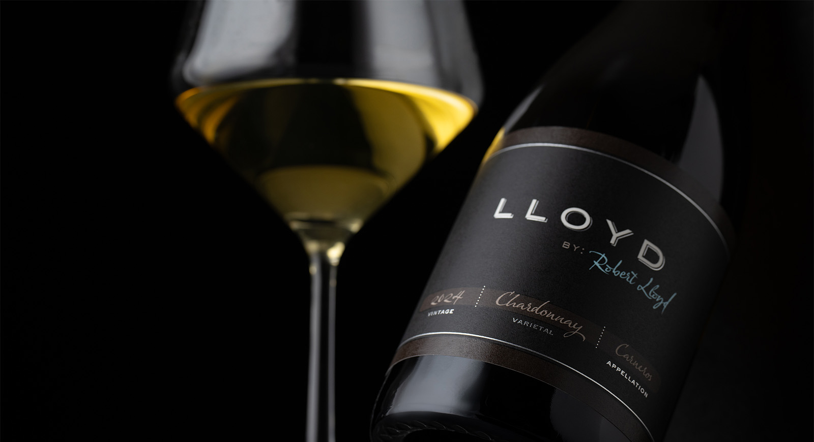 A close-up of a Lloyd by Robert Lloyd 2024 Carneros Chardonnay bottle beside a stemmed glass filled with golden white wine, both set against a dark background.