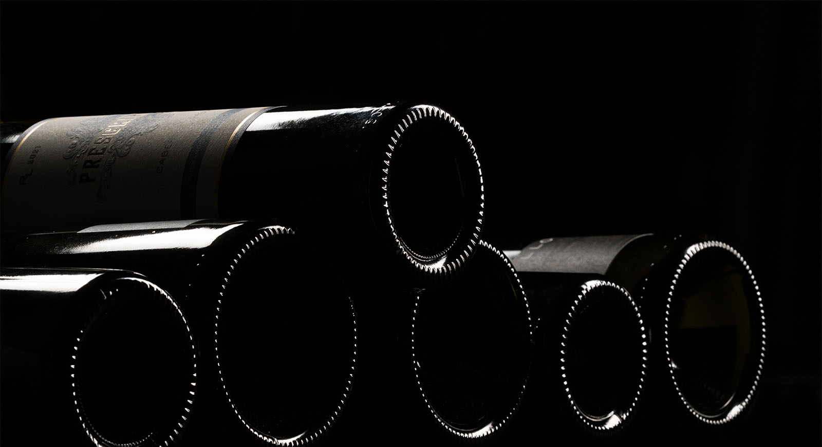 Wine bottles stacked horizontally in a dark cellar, their bases rimmed with light against a black background.