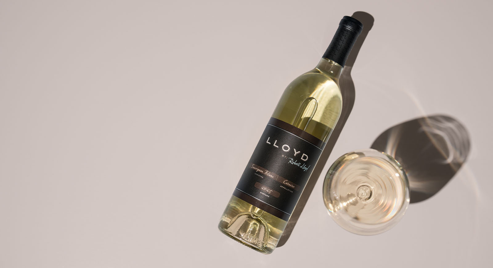 Bottle of 2025 Lloyd by Robert Lloyd Carneros Sauvignon Blanc lying on a pale cream surface beside a poured glass of white wine.